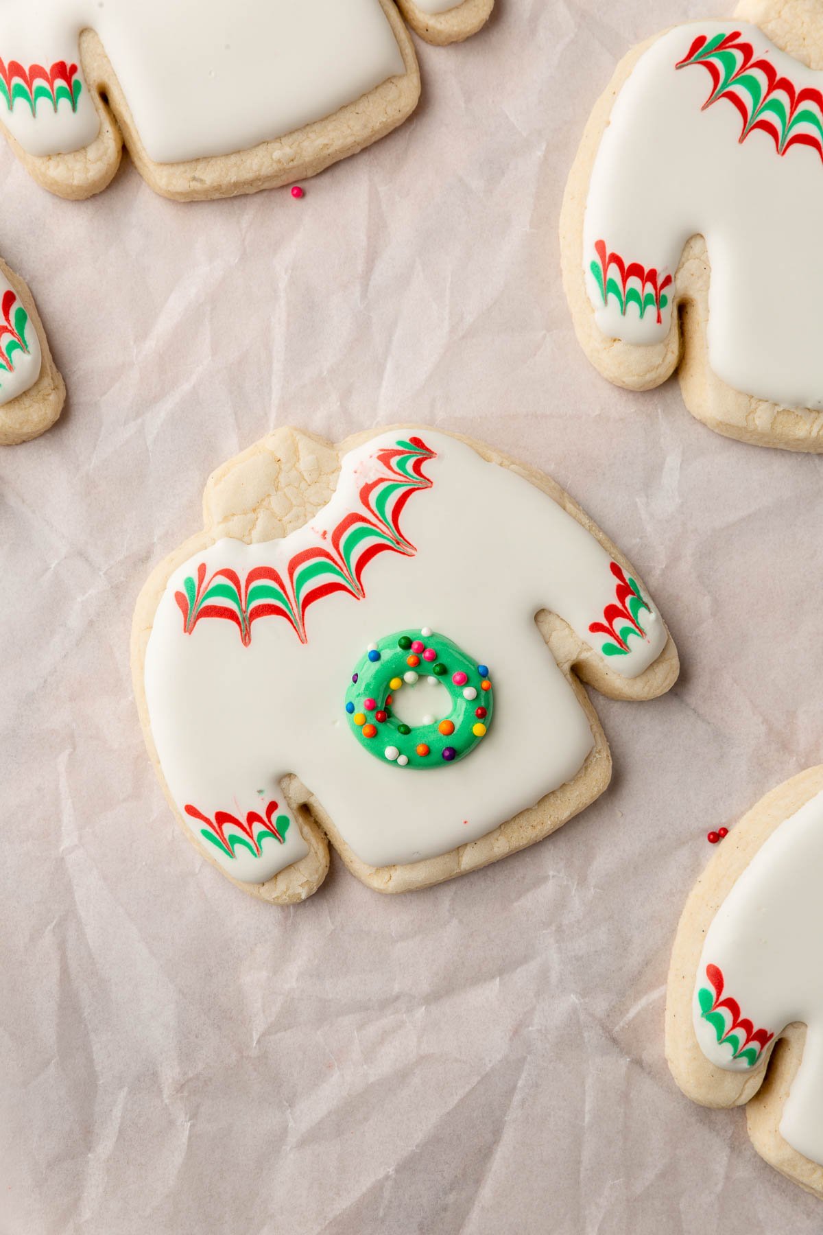 A gluten free ugly sweater sugar cookie with white royal icing and red and green chevron stripes with a green wreath in the center sprinkled with rainbow nonpareil sprinkles.