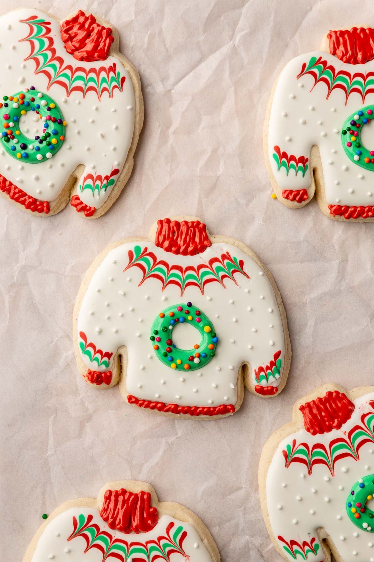 A gluten free ugly sweater sugar cookie with white royal icing and red and green chevron stripes with a green wreath in the center sprinkled with rainbow nonpareil sprinkles.