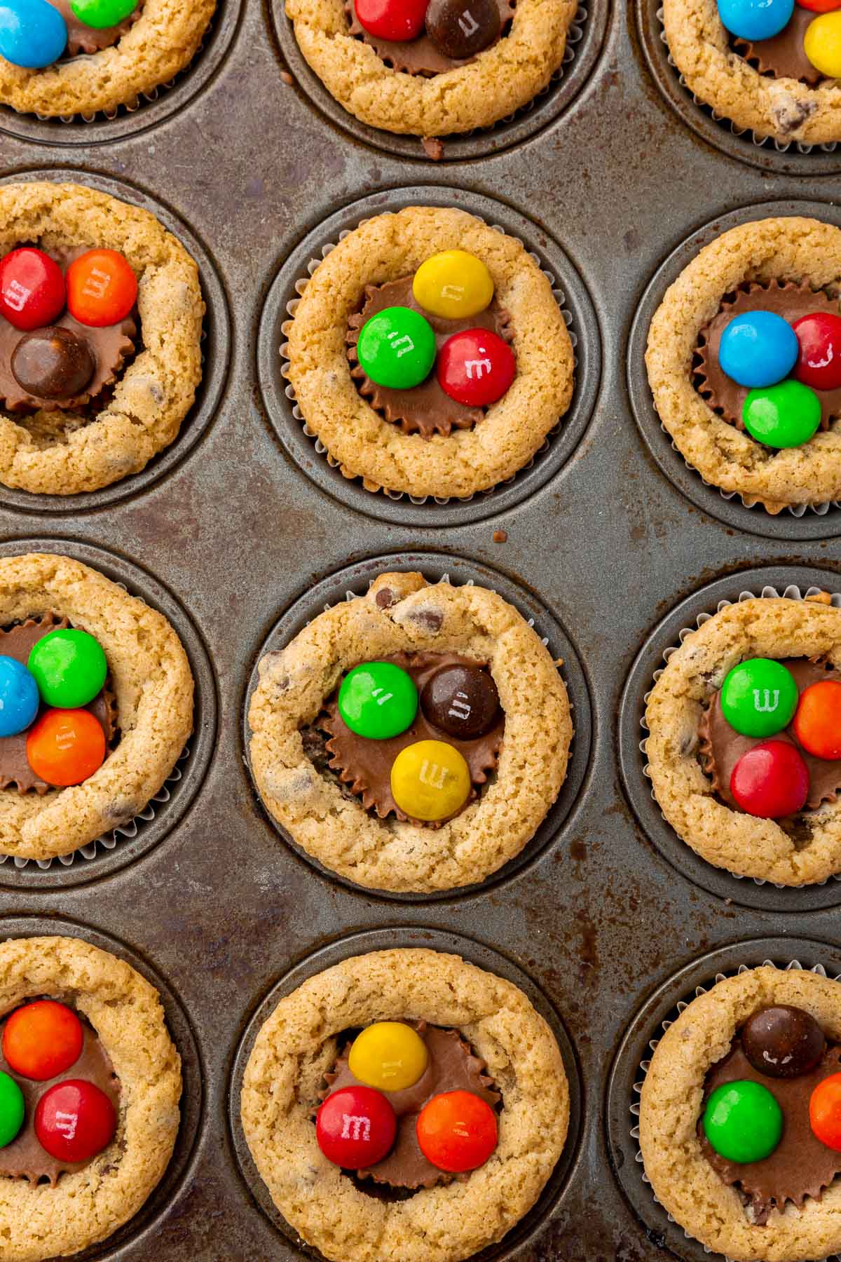 An overhead view of a mini muffin tin filled with chocolate chip cookie cups with a mini peanut butter cup in the center topped with 3 M&Ms each.