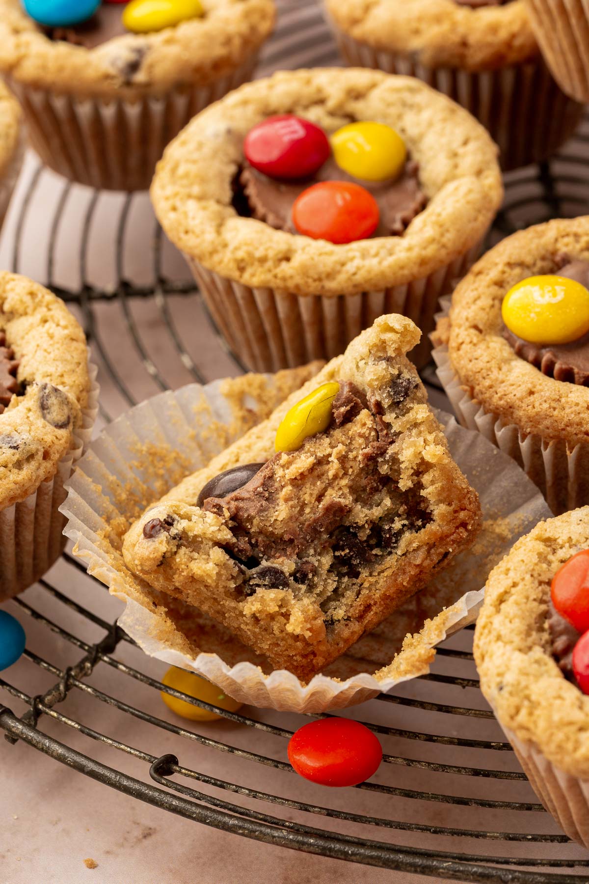 Multiple peanut butter cup cookies with M&Ms on a round wire cooling rack with the front cookie cup cut in half to see the inside.