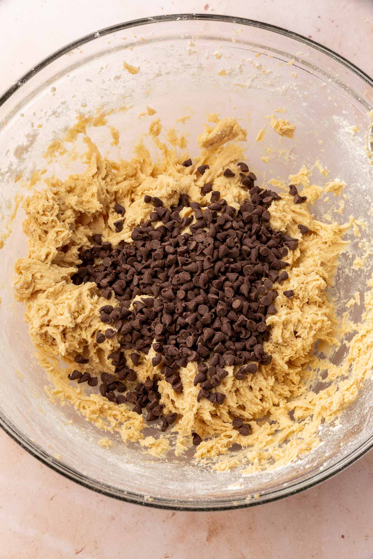 A glass mixing bowl of gluten-free cookie dough topped with a pile of mini chocolate chips before mixing together.