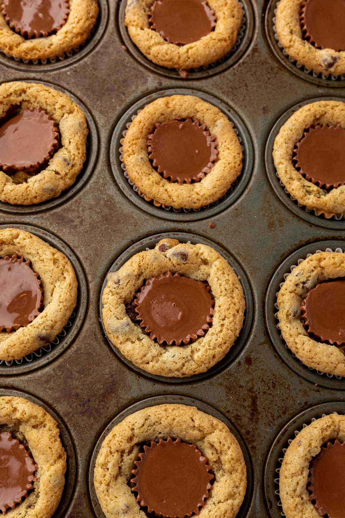 An overhead view of a mini muffin pan filed with chocolate chip cookie cups with mini peanut butter cups inserted in the center that have been melted.