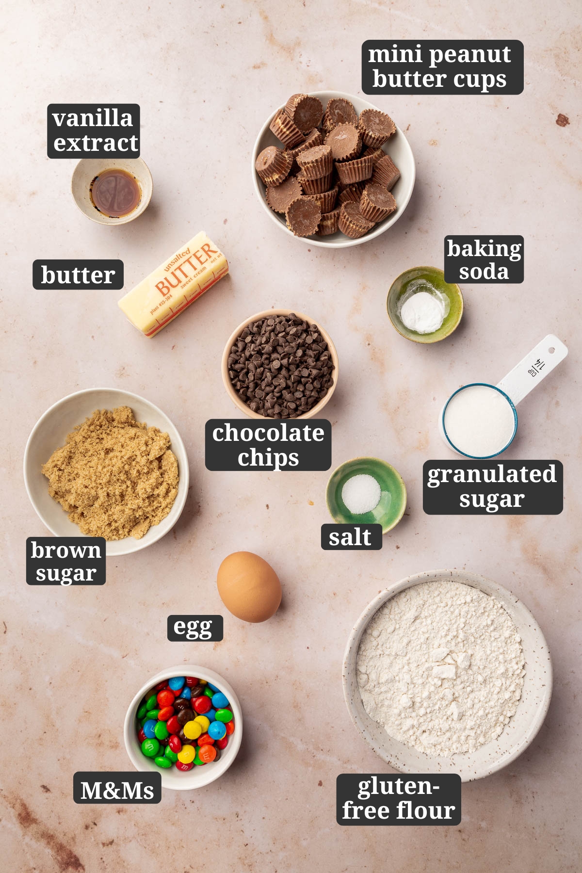 Ingredients in small bowls to make gluten-free peanut butter cup cookies with M&Ms, including mini peanut butter cups, vanilla extract, butter, chocolate chips, baking soda, brown sugar, egg, salt, granulated sugar, M&Ms and gluten-free flour with text overlays over each ingredient.