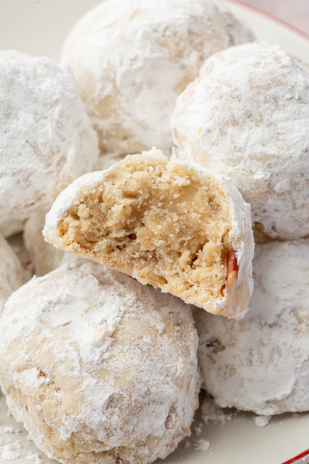 A mound of gf snowball cookies on a plate with the front snowball cookie cut in half to see the inside.