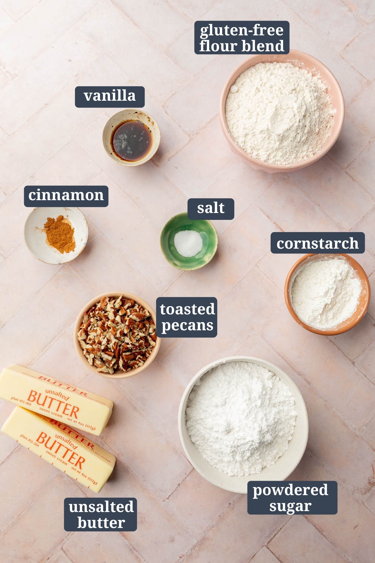 Ingredients in small bowls to make gluten free snowball cookies, including gluten-free flour blend, vanilla, cinnamon, salt, cornstarch, toasted pecans, powdered sugar, and unsalted butter with text overlays over each ingredient.