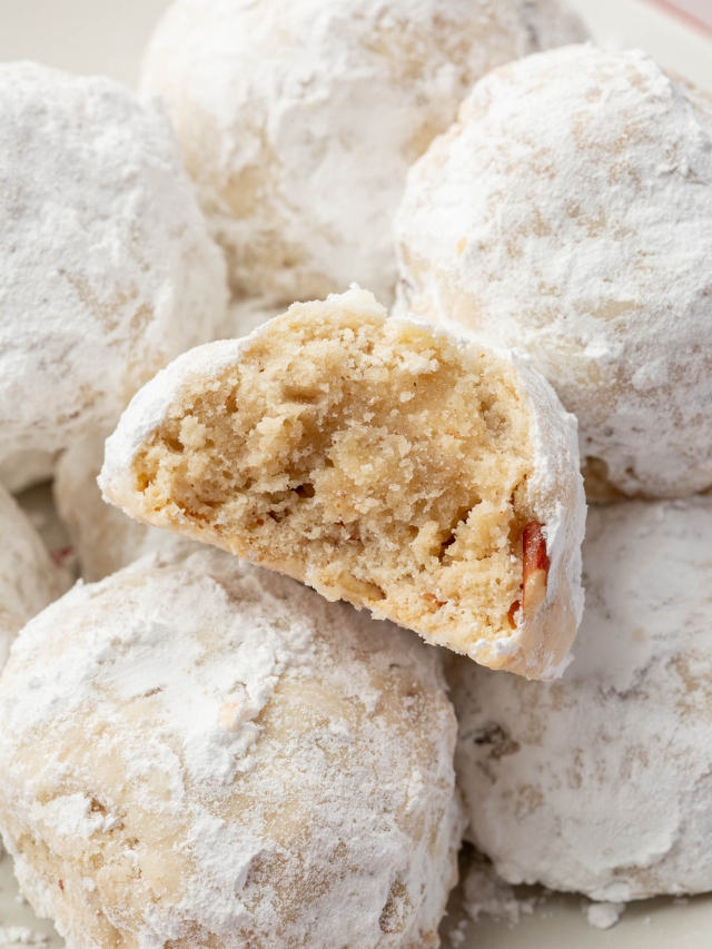 A mound of gf snowball cookies on a plate with the front snowball cookie cut in half to see the inside.