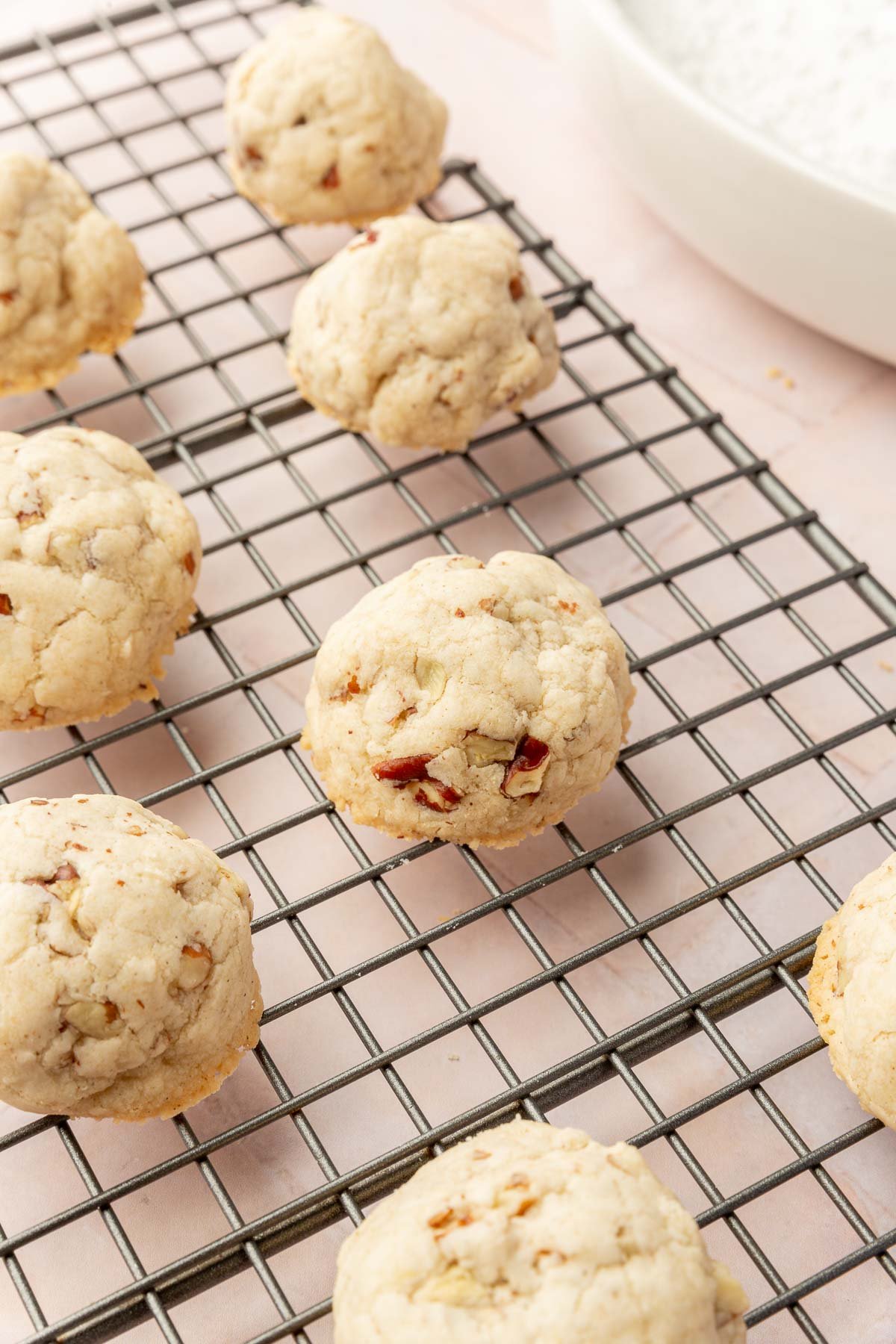 A wire cooling rack topped with gluten-free snowball cookies with pecans before rolling in powdered sugar.