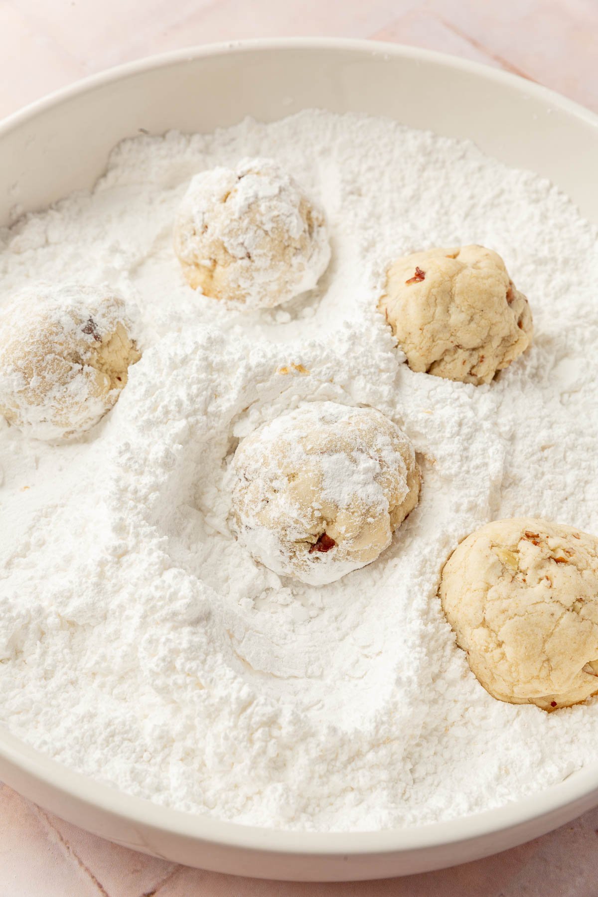 A low bowl with powdered sugar in it with five snowball cookies being dusted in powdered sugar.