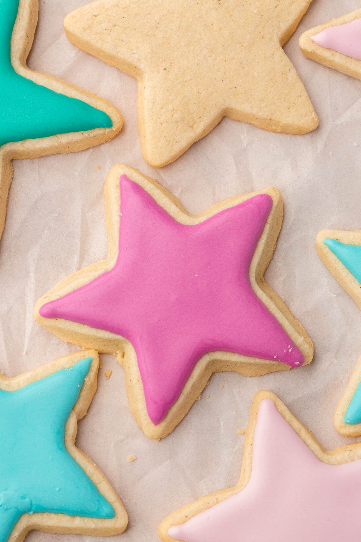 An overhead view of several gluten free sugar cookies ahped like stars and decorated with pink, blue, and green royal icing on a piece of crumpled parchment paper.
