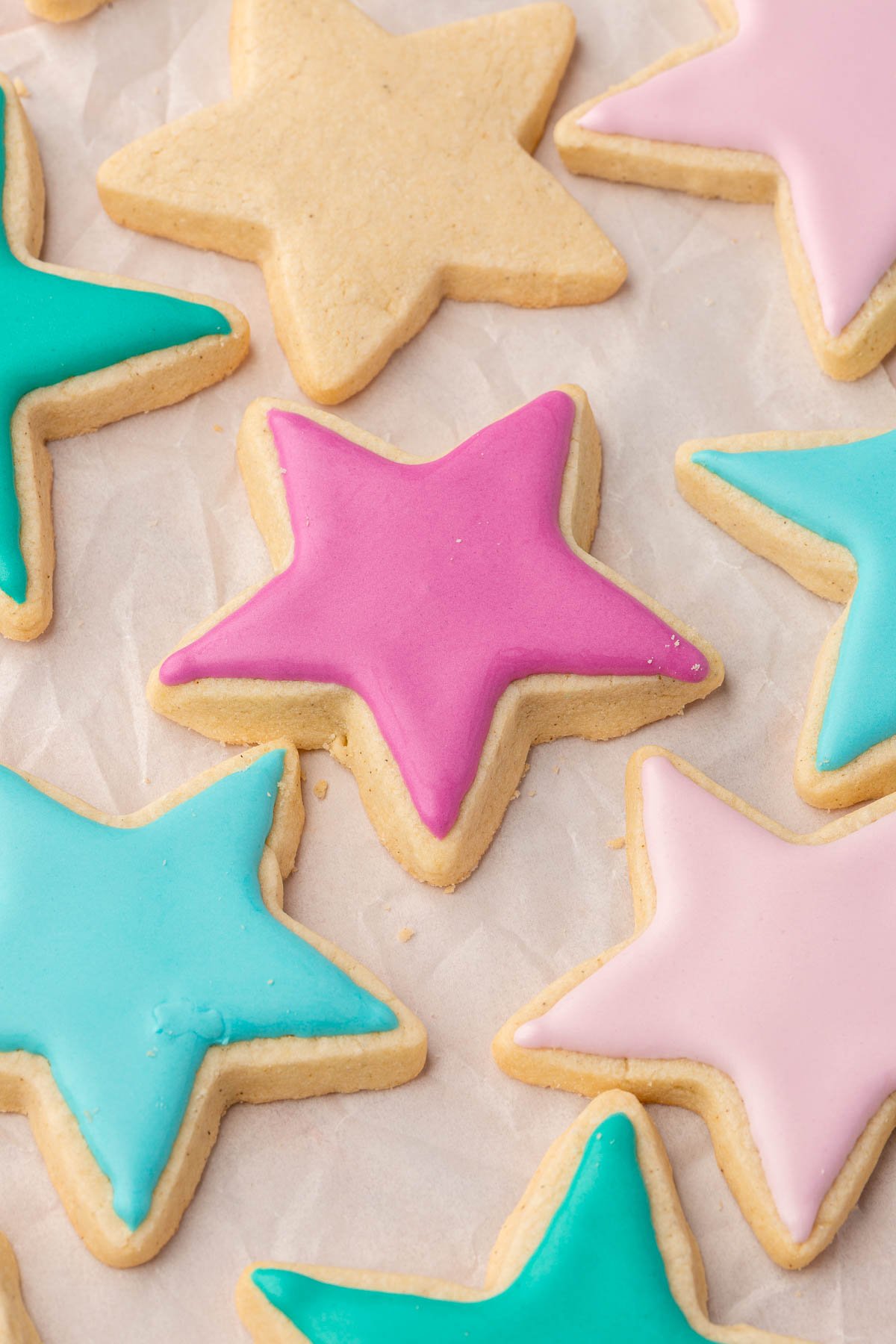 An overhead view of several gluten free sugar cookies ahped like stars and decorated with pink, blue, and green royal icing on a piece of crumpled parchment paper.