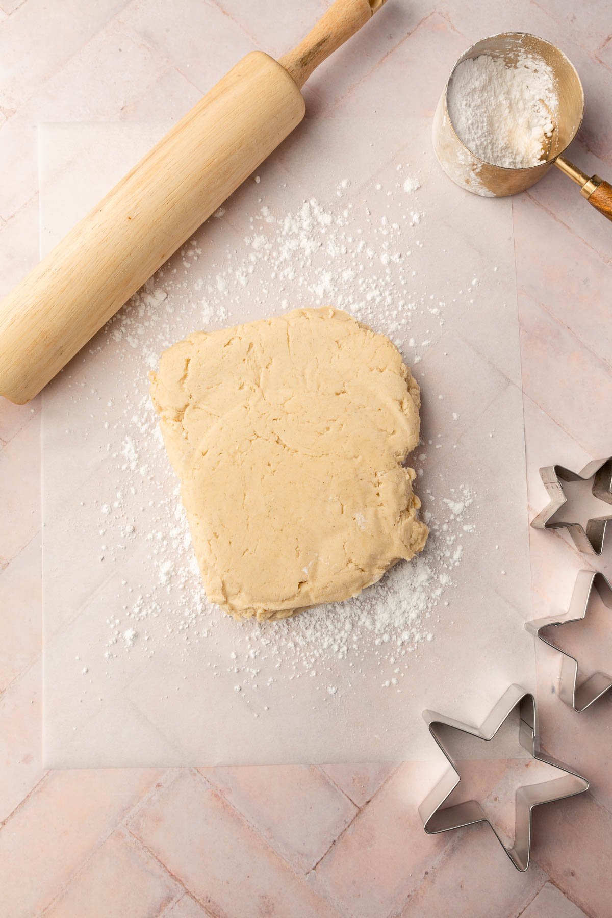 A slab of gluten free sugar cookie dough on a piece of parchment paper dusted with powdered sugar with a rolling pin, measuring cup of powdered sugar and star cookie cutters next to it.