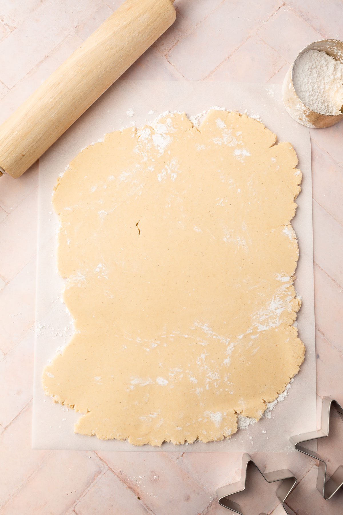 Gluten free sugar cookie dough rolled out onto a piece of parchment paper with a rolling pin, measuring cup of powdered sugar and star cookie cutters next to it.