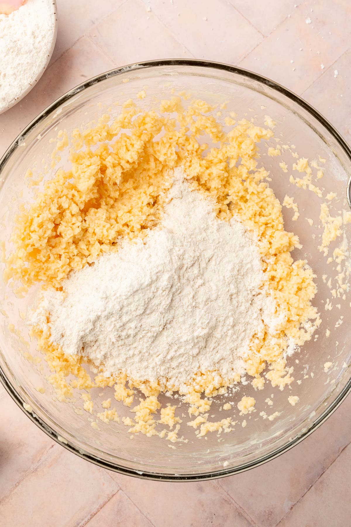 An overhead view of a butter, sugar and egg mixture topped with gluten-free flour in a glass mixing bowl.