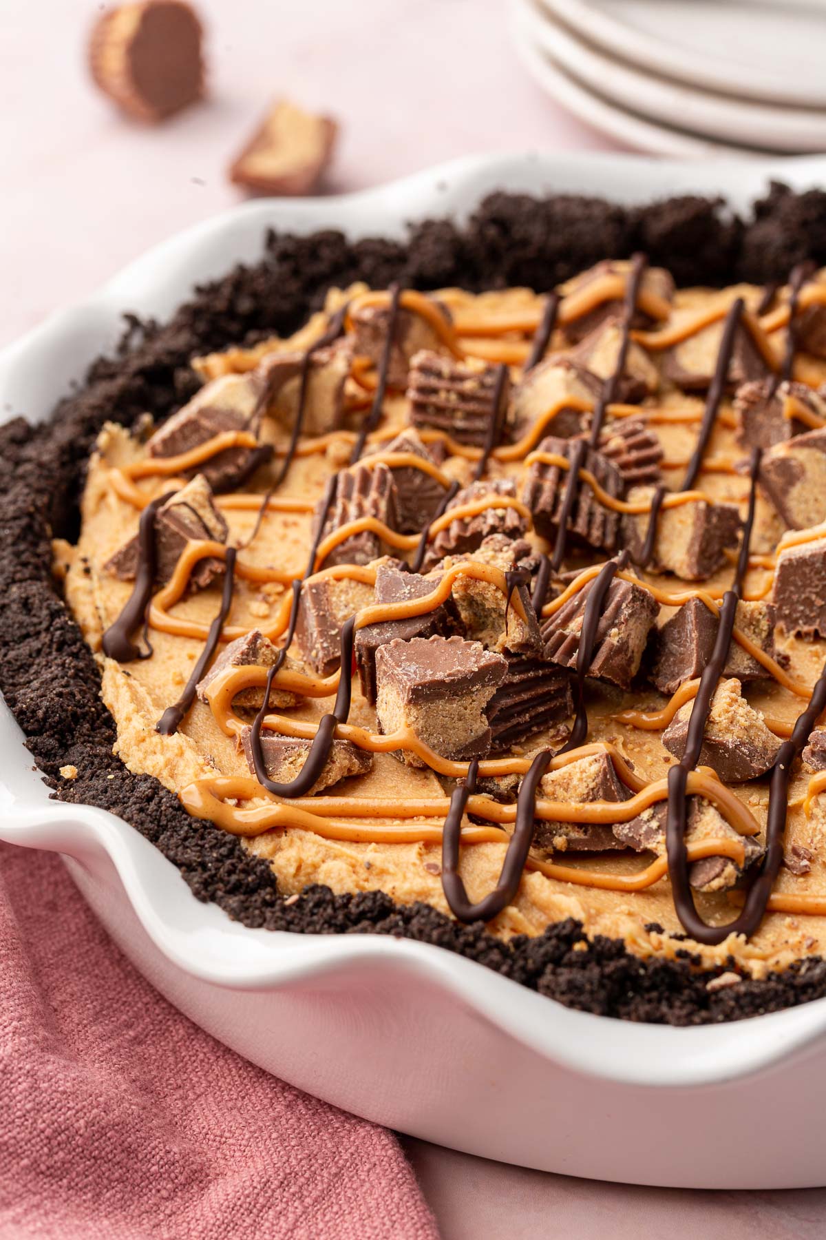 A whole gluten-free chocolate peanut butter pie with Oreo crust and topped with reese's peanut butter cups with a stack of plates in the background.