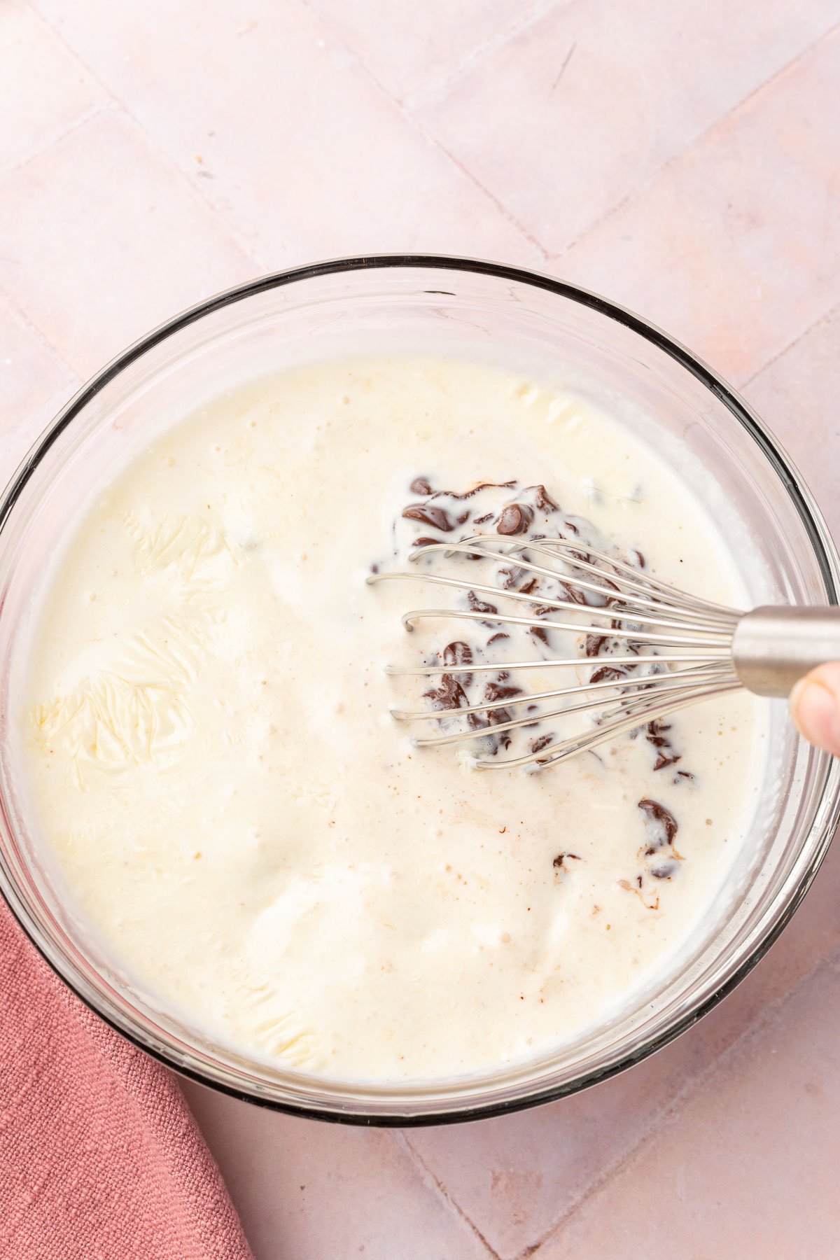 Hot heavy cream being whisked into chocolate chips in a glass mixing bowl to make ganache.