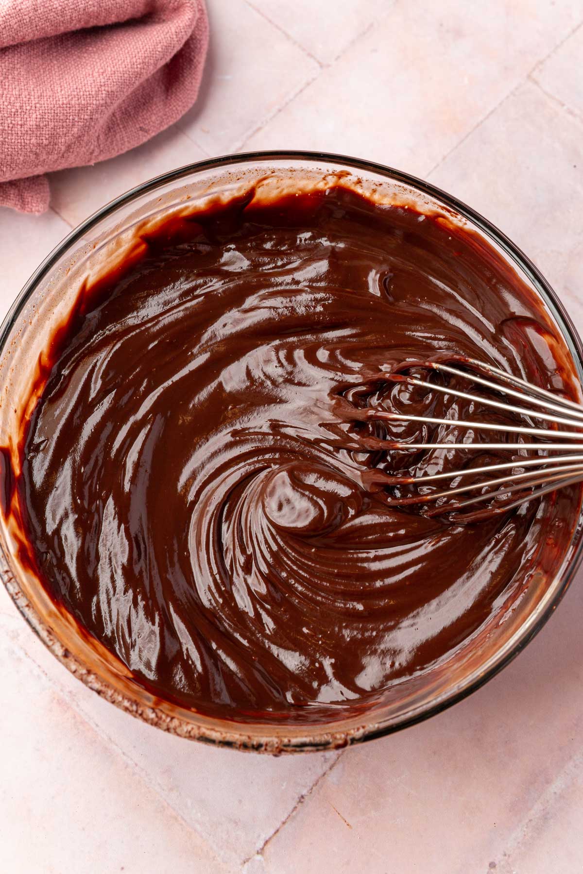 A glass mixing bowl with chocolate ganache and a wire whisk in it.