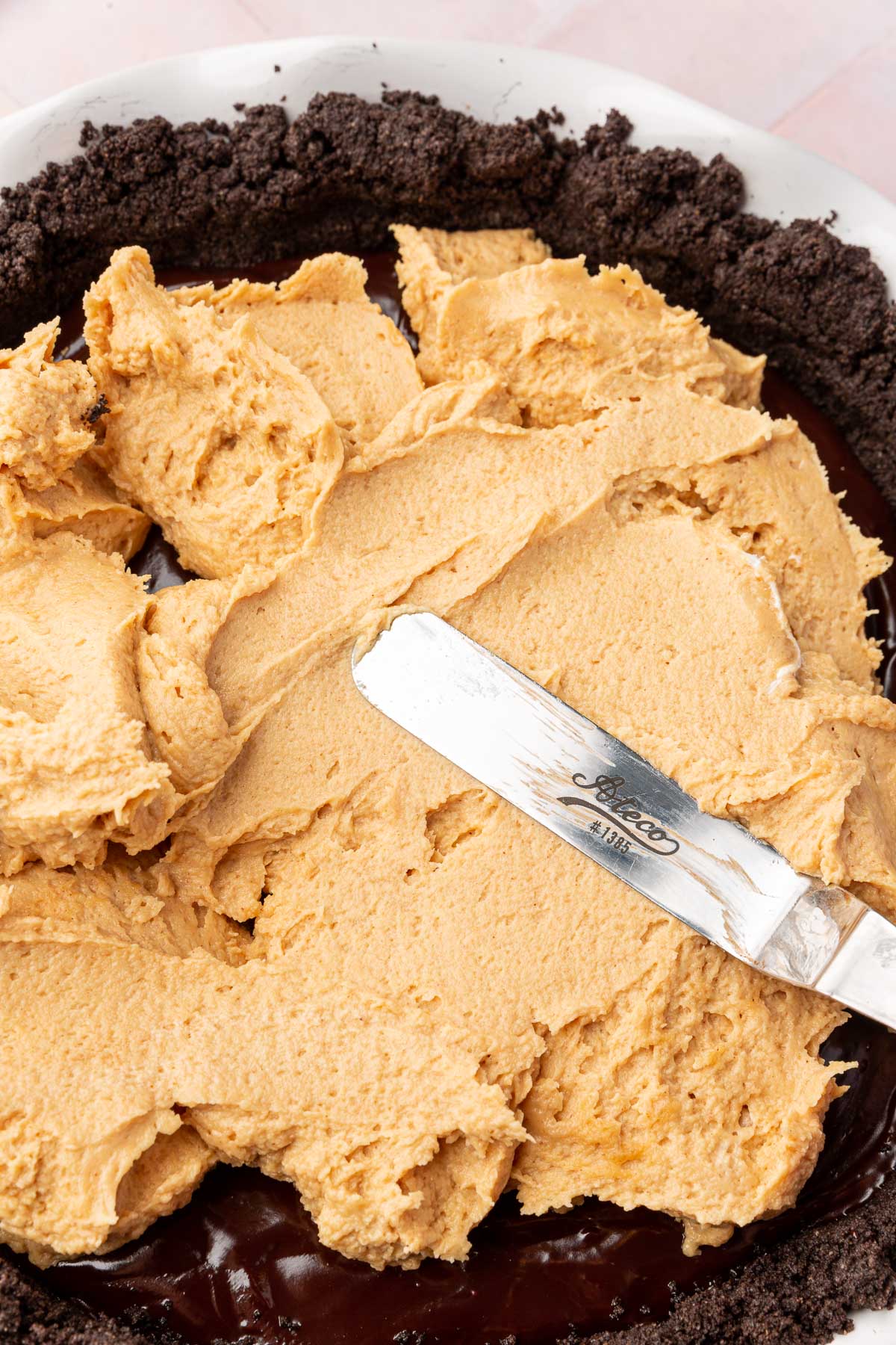 An offset spatula spreading a peanut butter filling over chocolate ganache on a gluten-free Oreo pie crust.