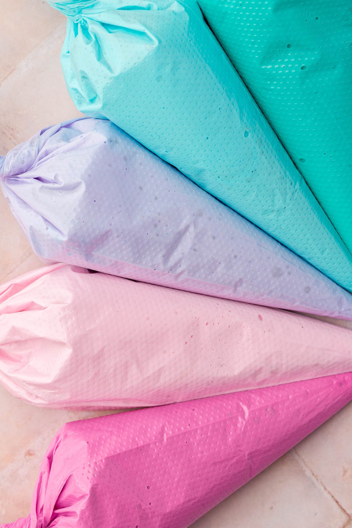 Five disposable pastry bags filled each with a different color of royal icing, teal, blue, purple, light pink and fuchsia.