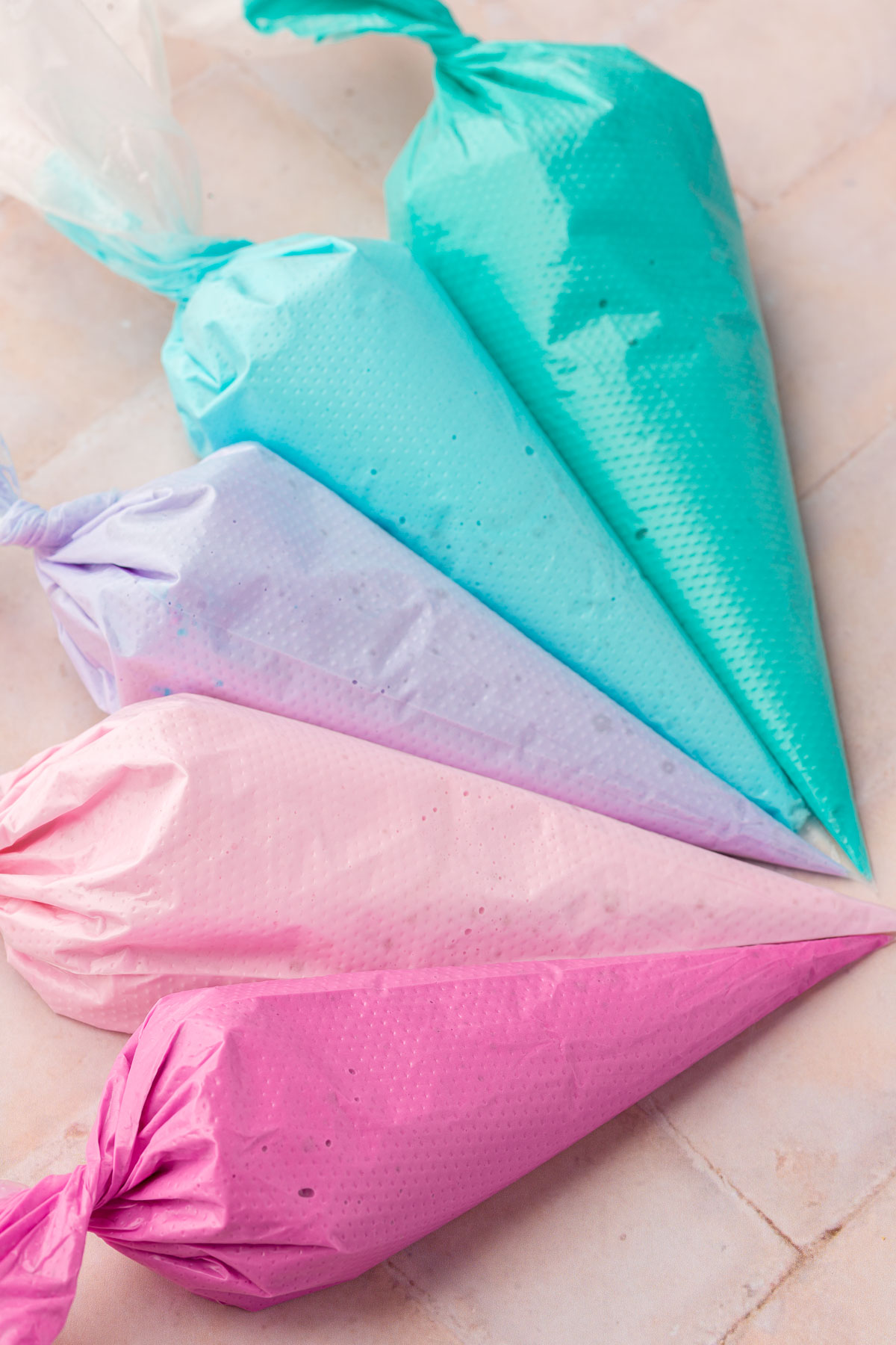 Five disposable pastry bags filled each with a different color of royal icing, teal, blue, purple, light pink and fuchsia.