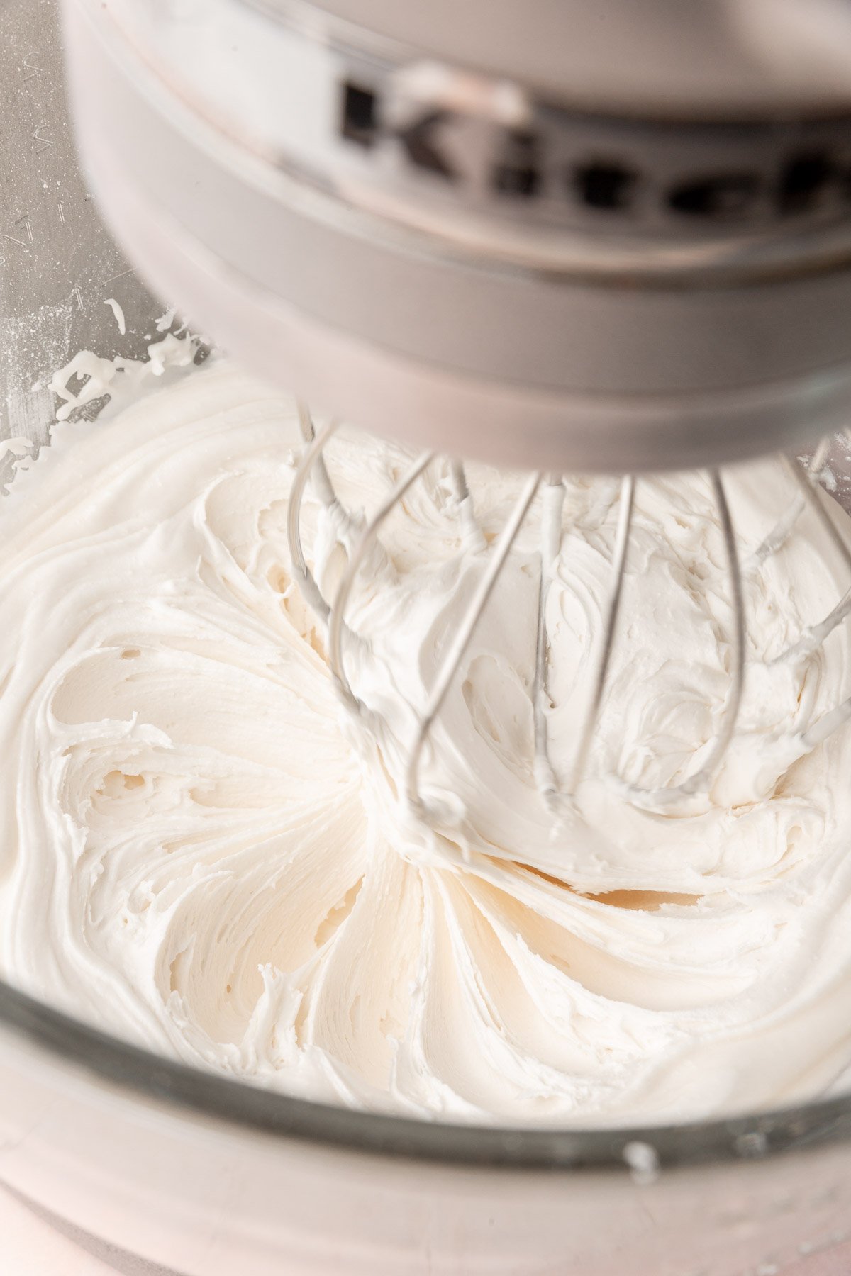 A stand mixer with a glass mixing bowl with the whisk attachment mixing a bowl of white royal icing.