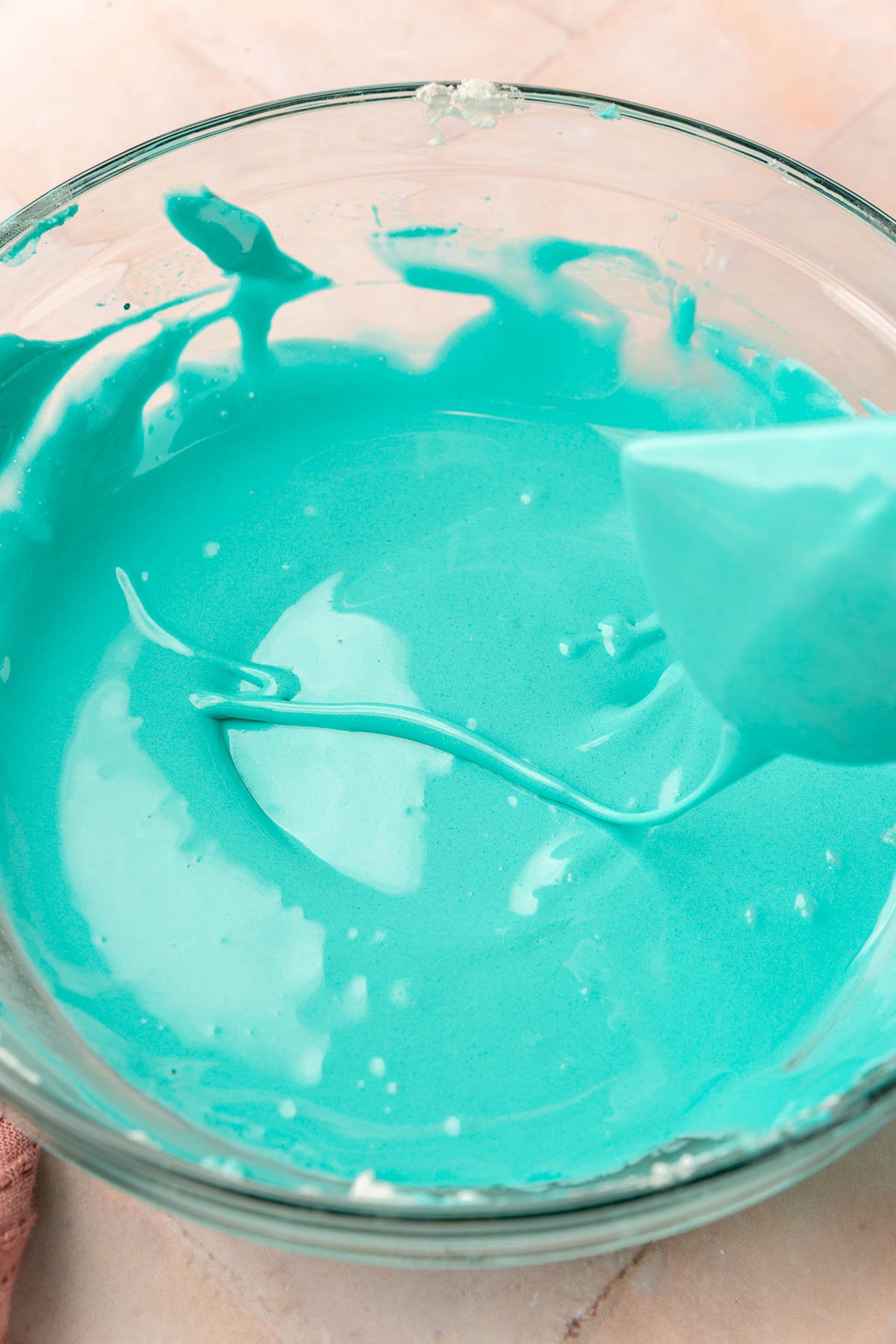 A glass mixing bowl of teal royal icing with a spatula illustrating the ribbon test for royal icing thickness.
