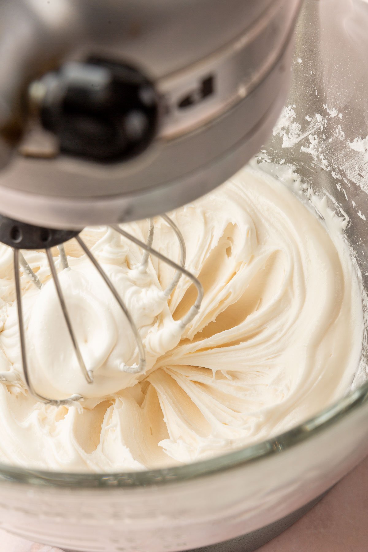 A stand mixer with a glass mixing bowl with the whisk attachment mixing a bowl of white royal icing.