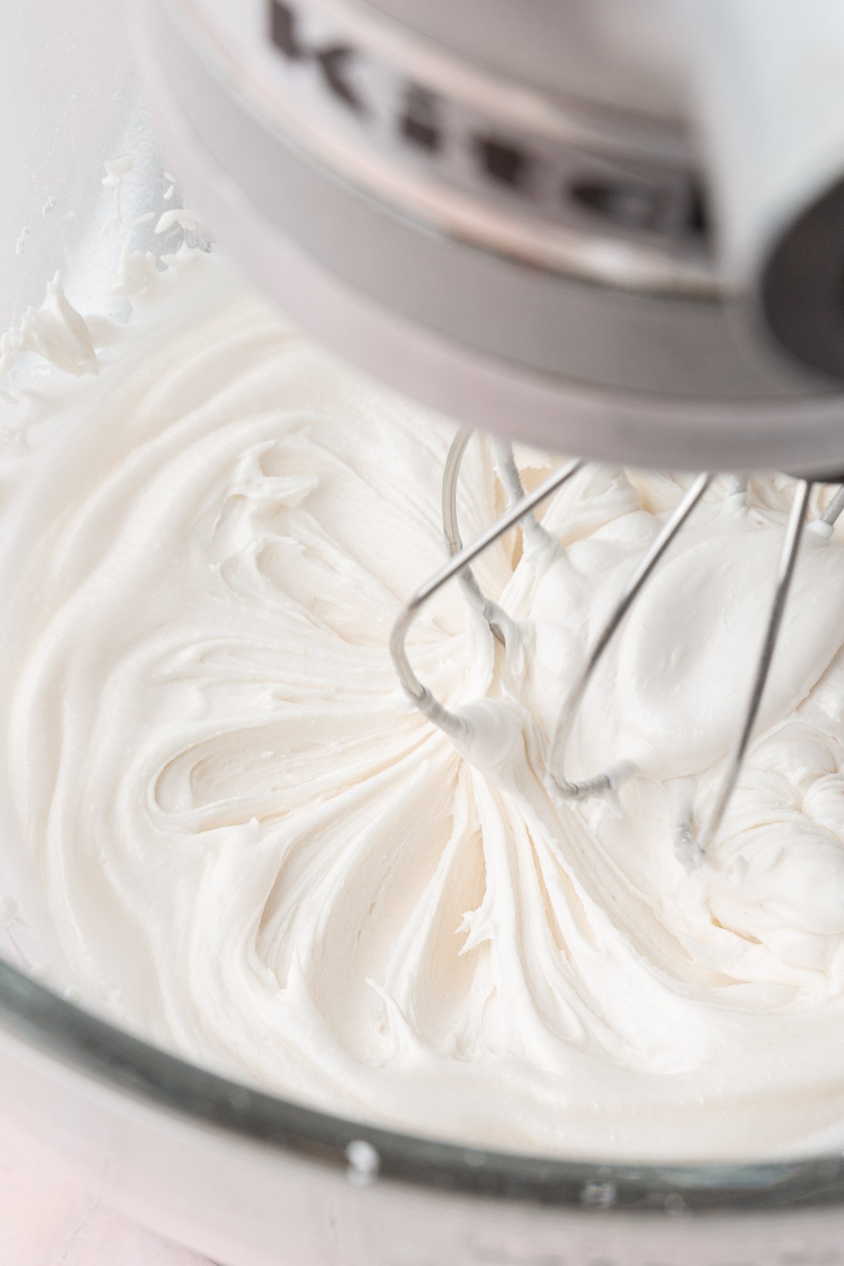A stand mixer with a glass mixing bowl with the whisk attachment mixing a bowl of white royal icing.