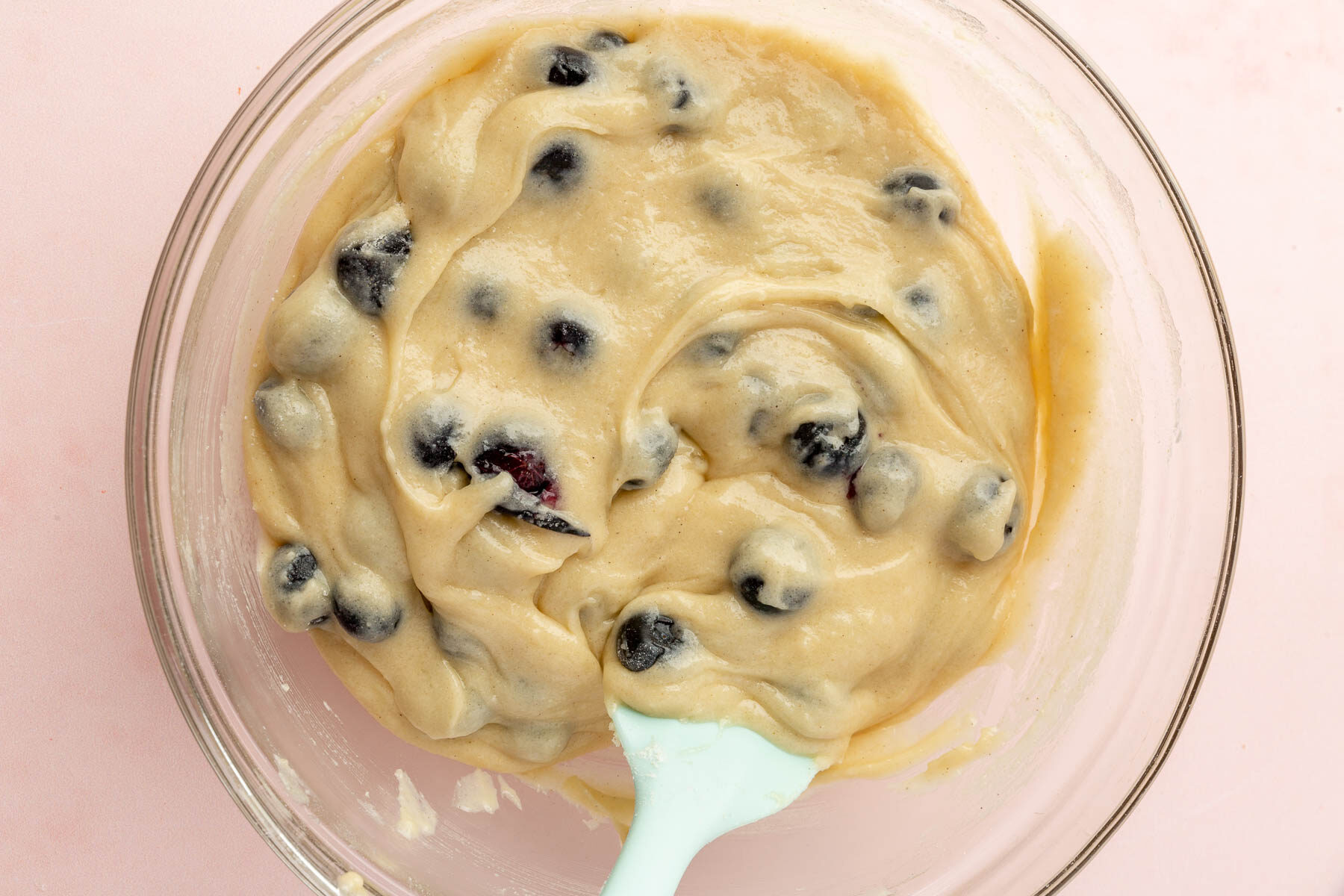 A glass mixing bowl with gluten-free blueberry muffin batter and a blue spatula in it.