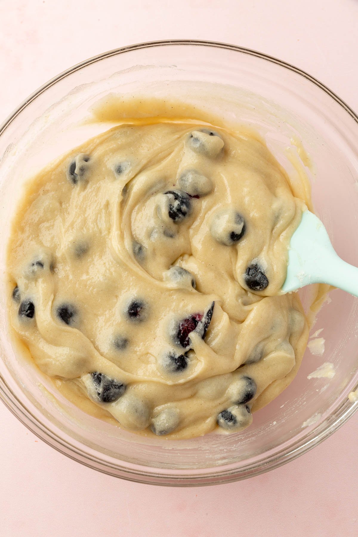 A glass mixing bowl with gluten-free blueberry muffin batter and a blue spatula in it.