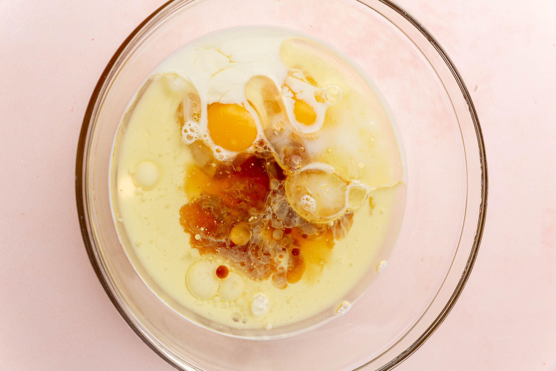 A glass mixing bowl with milk, oil, vanilla, and eggs before mixing together.