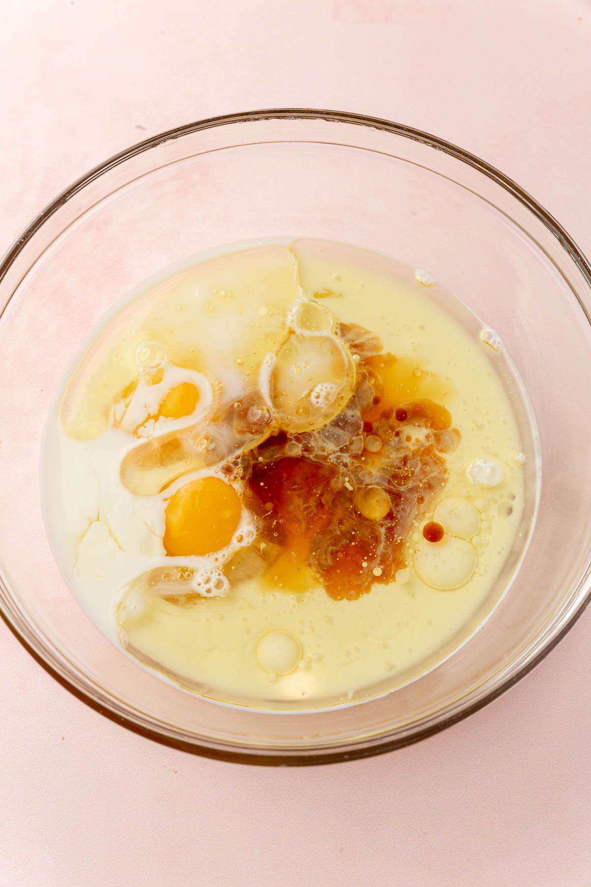 A glass mixing bowl with milk, oil, vanilla, and eggs before mixing together.