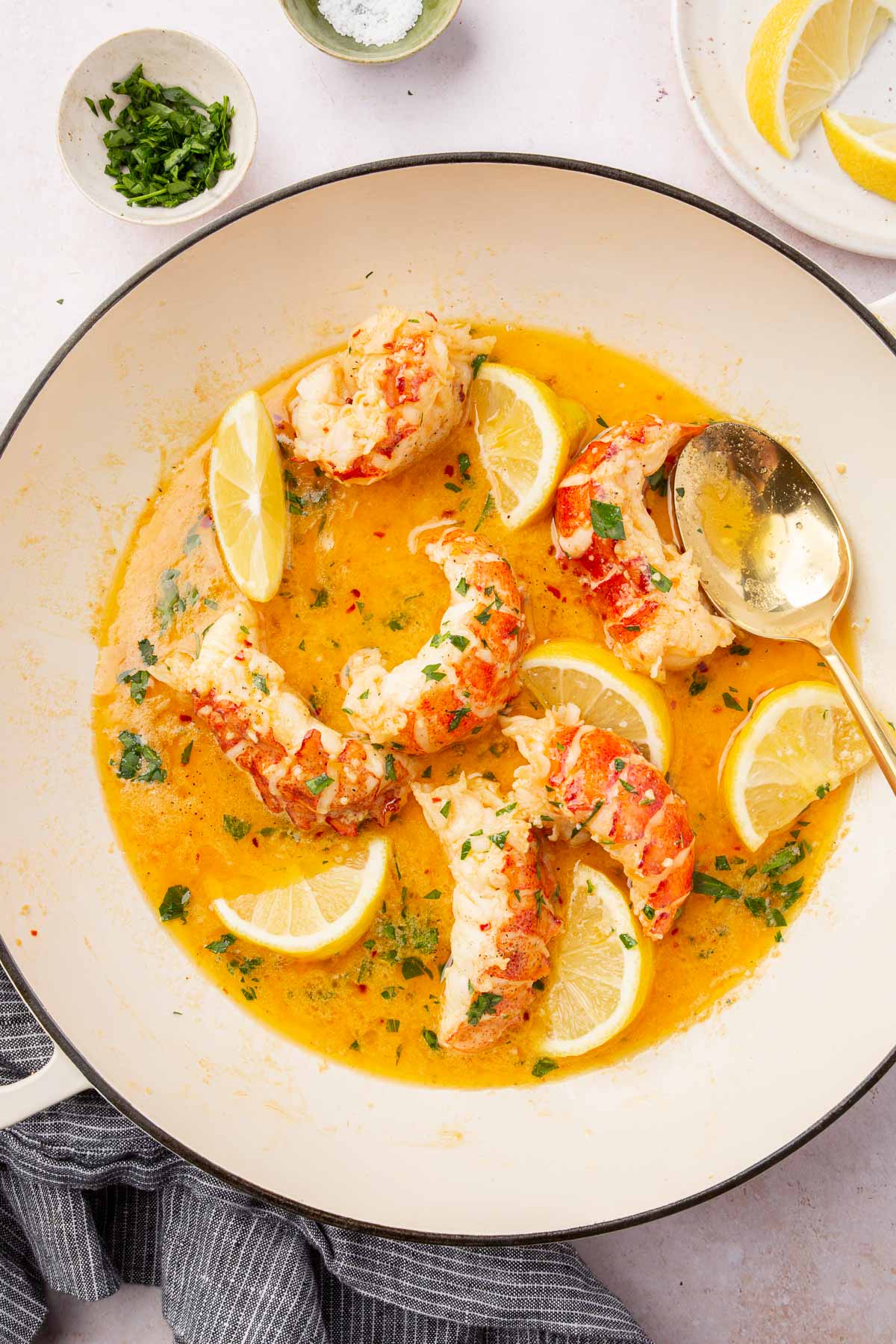 An overhead view of six lobster tails cooked in a butter garlic sauce in a braising pan with lemon wedges and fresh parsley.