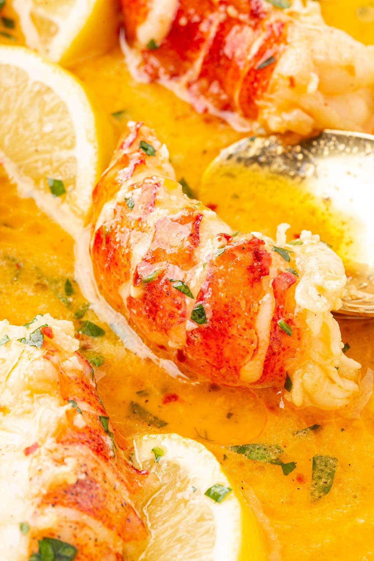 A close up of butter poached lobster tails in a pool of beurre monté poaching liquid with parsley, lemon wedges, and a spoon.