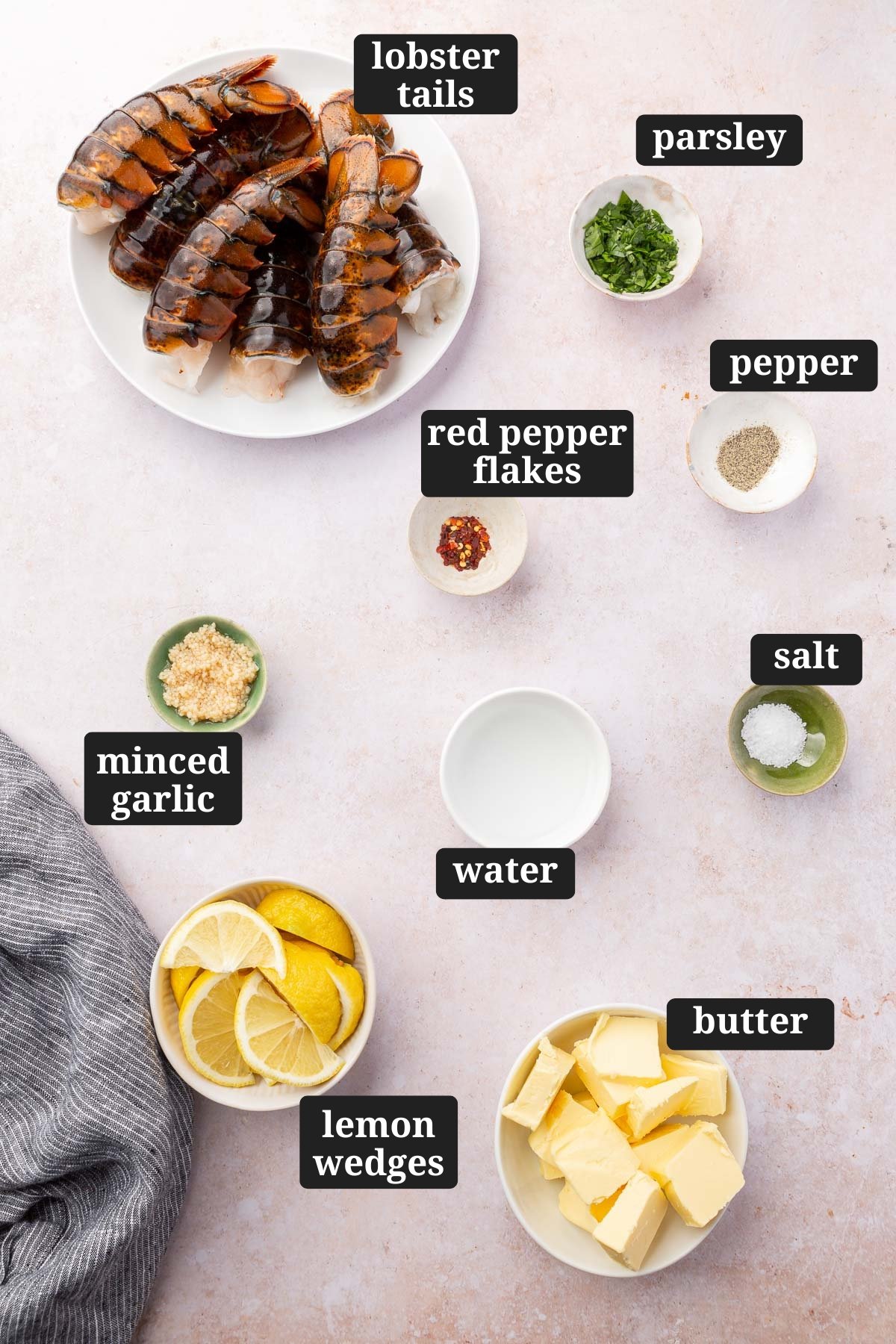 An overhead view of ingredients in small bowls to make butter poached lobster tails, including raw lobster tails, parsley, pepper, red pepper flakes, minced garlic, water, salt, lemon wedges and butter with text overlays over each ingredient.