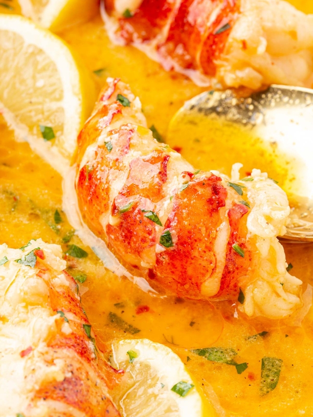 A close up of butter poached lobster tails in a pool of beurre monté poaching liquid with parsley, lemon wedges, and a spoon.