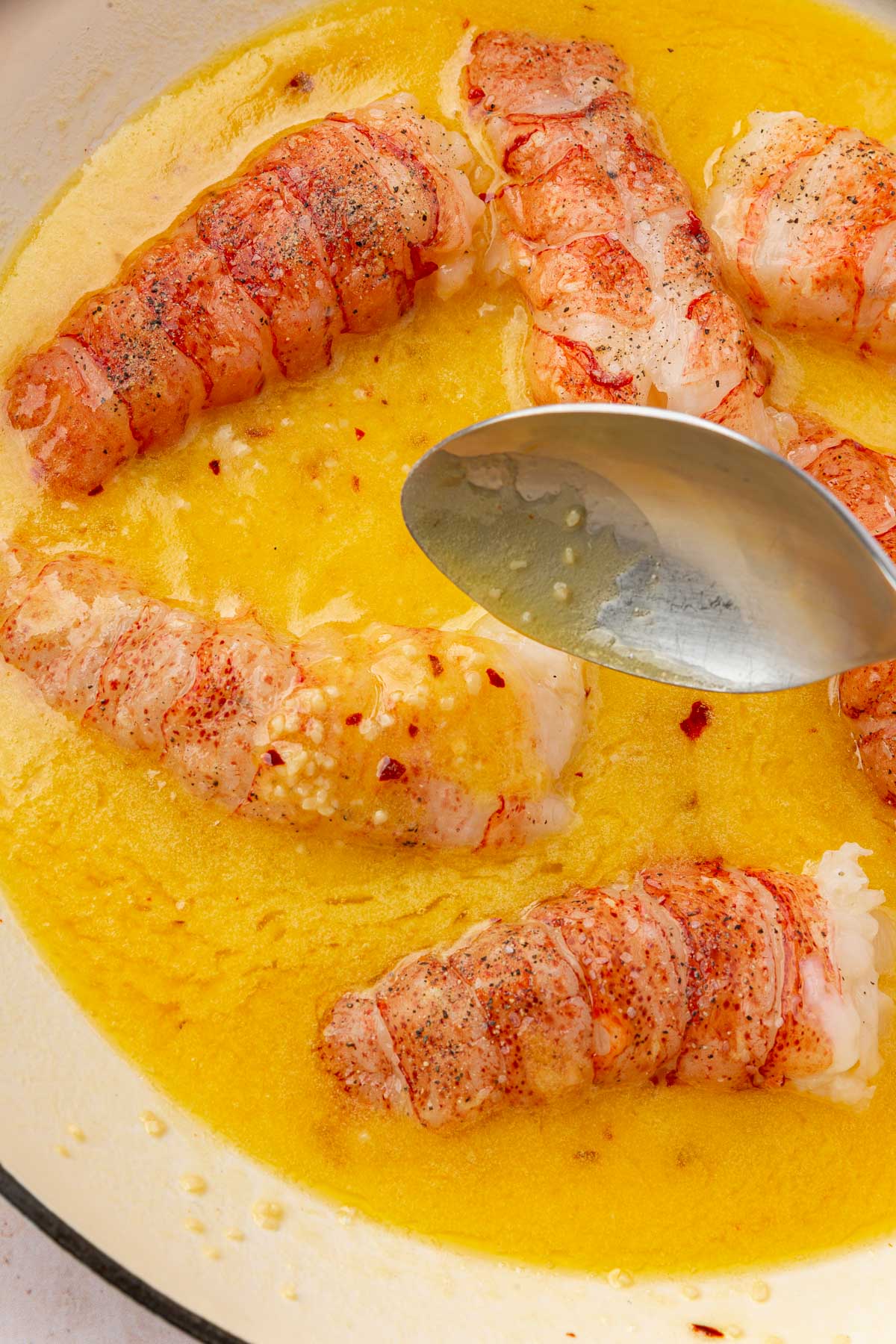 A silver spoon pouring butter garlic sauce over raw lobster tails in a braising pan to make butter poached lobster.