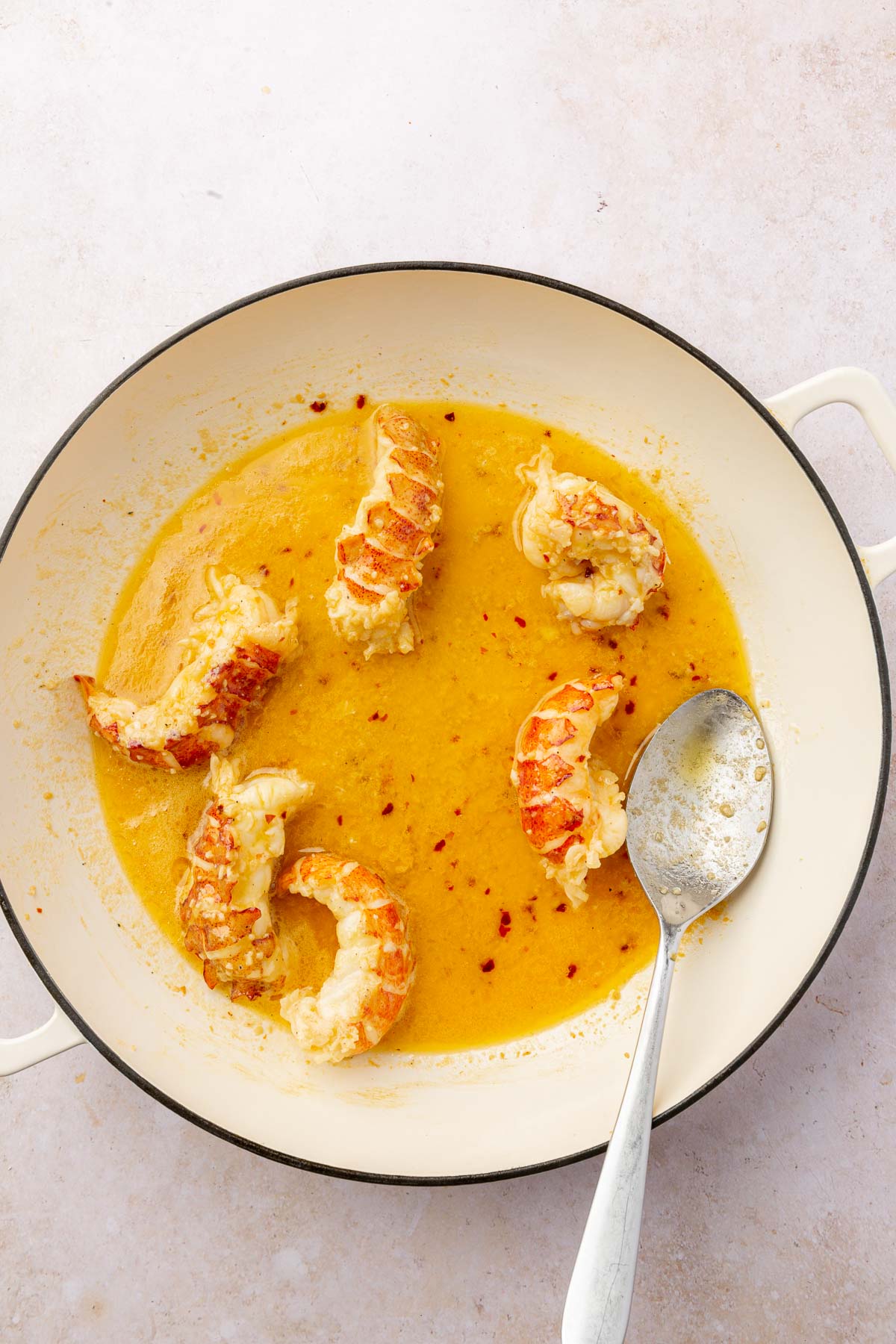 An overhead view of six lobster tails in a butter poaching liquid in a braising pan with a spoon.