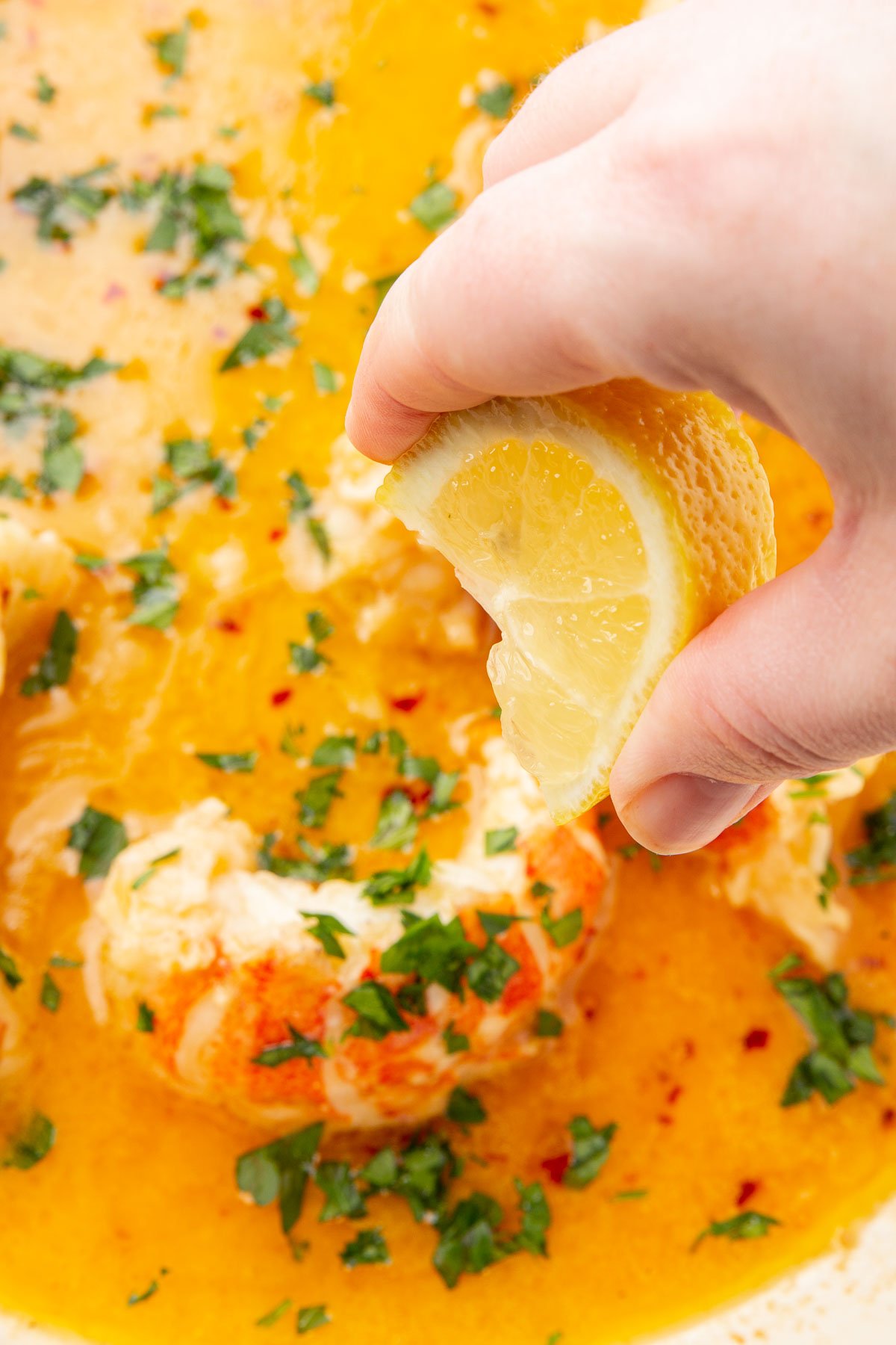 A hand squeezing a lemon wedge over butter poached lobster tails in a beurre monté with parsley.