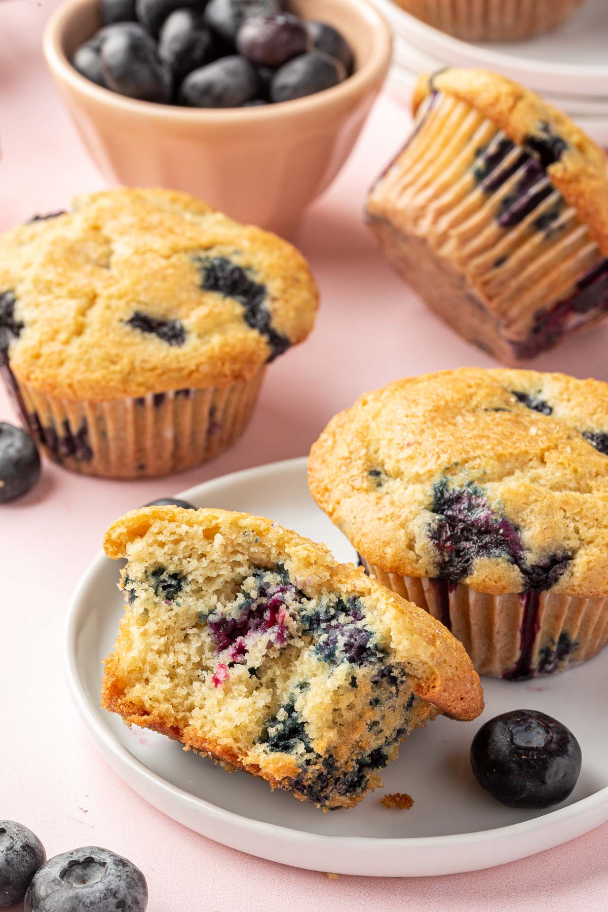 A gluten free blueberry muffin cut in half on a small plate with additional muffins and fresh blueberries in the background.