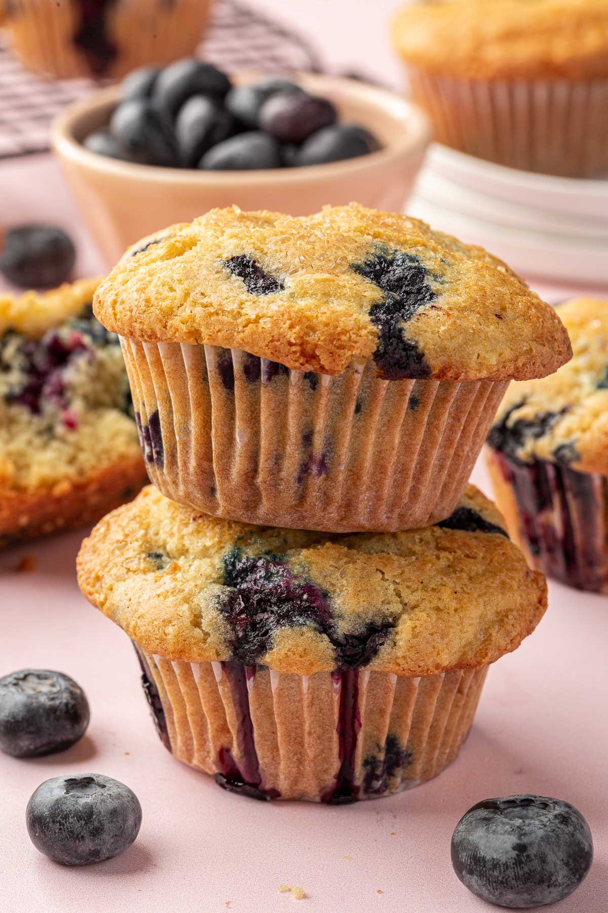 Two gluten free blueberry muffins stacked on top of each other with additional blueberry muffins and fresh blueberries in the background.