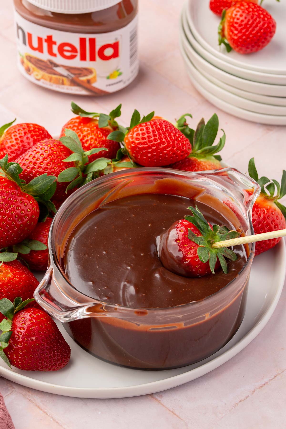 A ramekin of nutella ganache with a skewered strawberry dipped into it on a plate of whole strawberries with a pink linen napkin next to it.