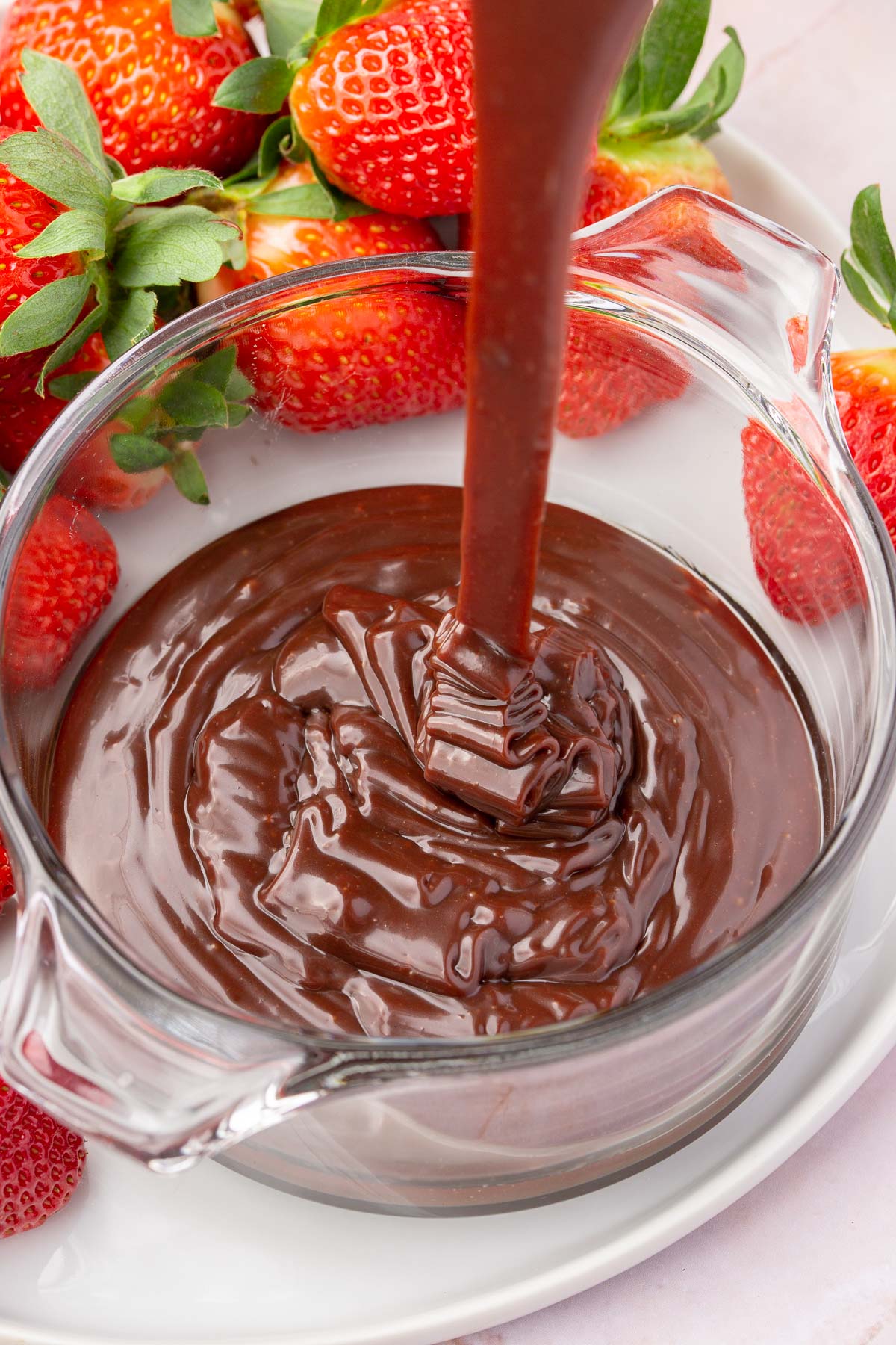 Nutella ganache being drizzled into a glass ramekin on a plate of fresh strawberries.