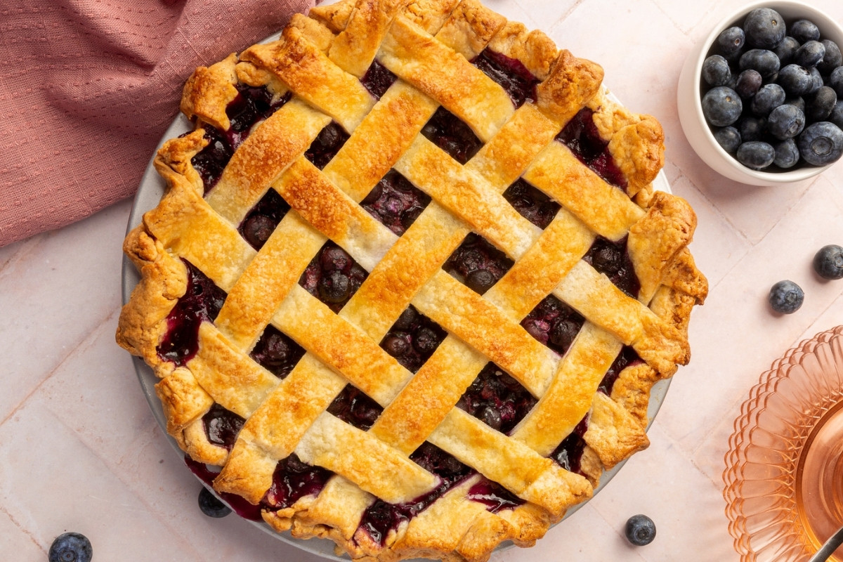 A freshly baked blueberry pie with a golden-brown lattice crust sits on a light surface, surrounded by gluten-free blueberry muffins, a few scattered blueberries, a pink napkin, and a bowl of fresh blueberries.