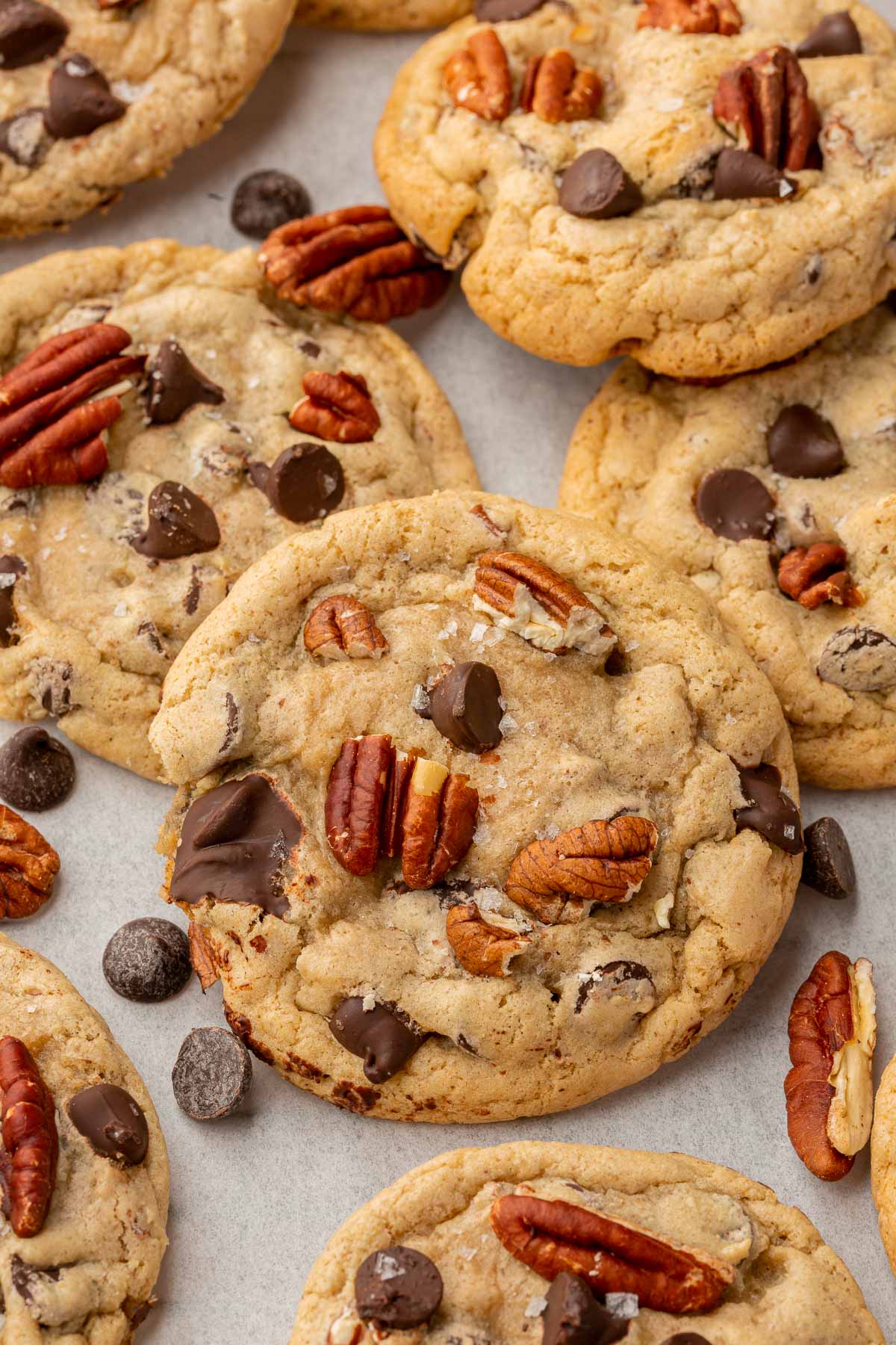 A close up of gluten-free chocolate chip pecan cookies layered on top of each other on a piece of parchment paper with toasted pecans and dark chocolate chips scattered around.