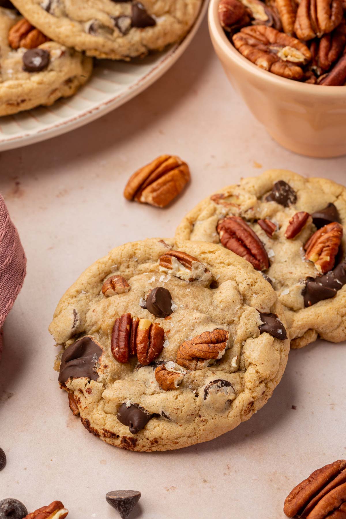 Two gluten free pecan chocolate chip cookies layered on top of each other on a pink table with additional gluten free cookies and toasted pecans in the background.