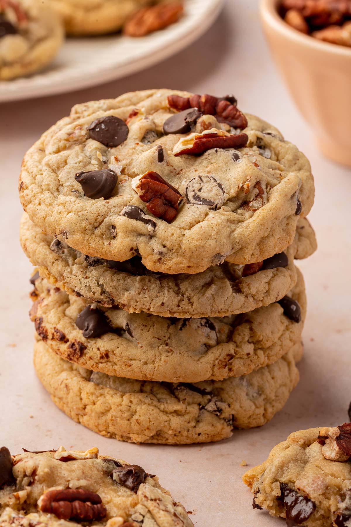 A stack of four gluten free pecan chocolate chip cookies on a pink table with additional cookies and toasted pecans in the background.