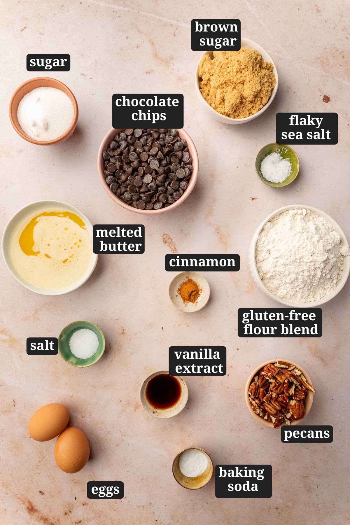 Ingredients in small bowls to make gluten free pecan chocolate chip cookies, including brown sugar, sugar, chocolate chips, flaky sea salt, melted butter, cinnamon, gluten-free flour blend, vanilla, salt, pecans, baking soda, and eggs with text overlays over each ingredient.
