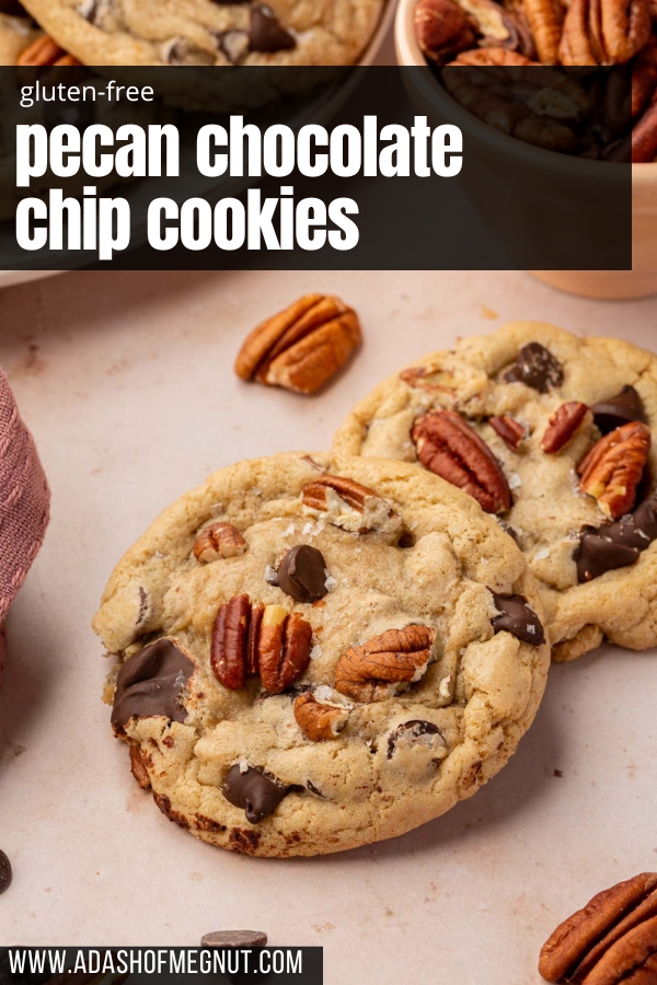 Two gluten free pecan chocolate chip cookies layered on top of each other on a pink table with additional gluten free cookies and toasted pecans in the background.