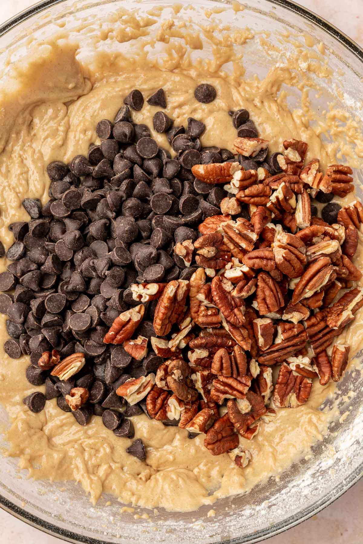A close up of gluten free chocolate chip cookie dough topped with a pile of dark chocolate chips and toasted pecans before mixing together.