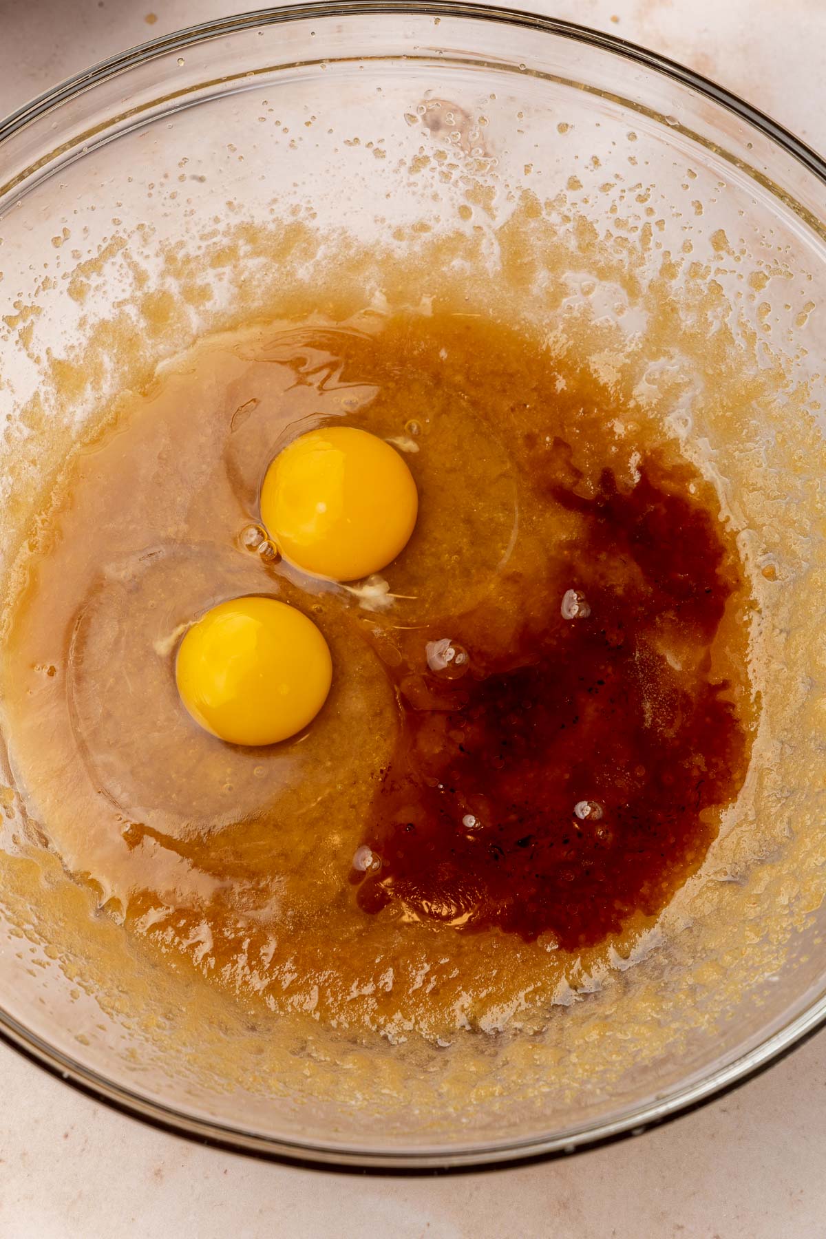 A glass mixing bowl with a mixture of melted butter, brown sugar and granulated sugar topped with pure vanilla extract and two eggs.